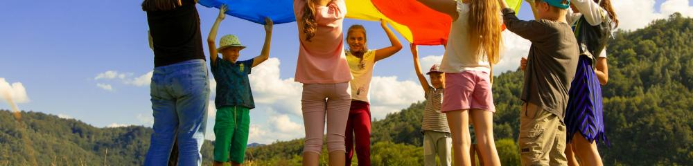 Children with a play parachute in a field under a blue sky with clouds - by Artem Kniaz via Unsplash       